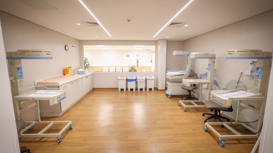 Descrição para fins de acessibilidade: Sala do centro obstétrico de um hospital, ampla, com piso amadeirado e iluminação em linhas de LED no teto. À direita, três estações de cuidado neonatal com berços aquecidos transparentes sobre rodízios e cadeiras giratórias. À esquerda, outra estação semelhante ao lado de uma bancada com pia, dispensers e um relógio de parede. No fundo, há uma janela interna para o corredor e cinco lixeiras com tampas coloridas para separação de resíduos. O ambiente está vazio, organizado e pronto para uso.
