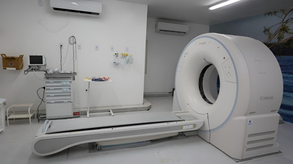 Ambiente clínico branco, bem iluminado, destinado a exames de imagem. À direita está o grande tomógrafo computadorizado da Canon, circular, com anel amplo e mesa motorizada alinhada ao centro do equipamento. À esquerda, um carrinho de procedimentos com monitor, gavetas e cabos; acima dele, tomadas e pontos de gases medicinais na parede. Há também uma mesinha com materiais e dois aparelhos de ar-condicionado do tipo split instalados no alto das paredes. O piso é cinza claro e, ao fundo, parte da parede tem um mural com água azul e ramos de bambu. Não há pessoas na cena; o espaço está organizado e pronto para uso.