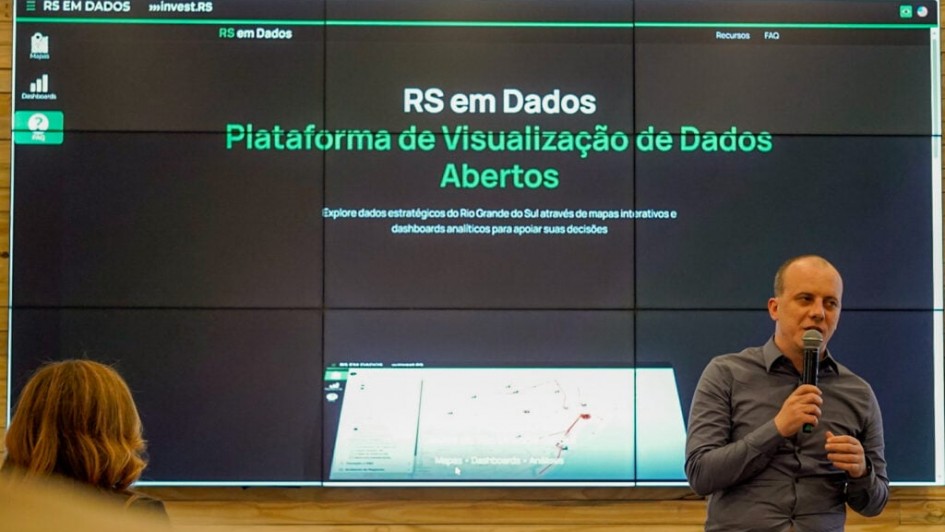 A imagem mostra uma apresentação em um ambiente interno, provavelmente um auditório ou sala de conferências. No centro da imagem, há uma grande tela dividida em vários painéis exibindo a plataforma "RS em Dados", com o texto:
"RS em Dados – Plataforma de Visualização de Dados Abertos. Explore dados estratégicos do Rio Grande do Sul através de mapas interativos e dashboards analíticos para apoiar suas decisões."
À direita da imagem, está um homem careca, vestindo camisa cinza, segurando um microfone enquanto fala. Ele parece estar apresentando ou explicando a plataforma. À esquerda, de costas, está uma mulher assistindo à apresentação.
O ambiente sugere uma ocasião formal de lançamento ou demonstração de um sistema digital governamental. A interface da plataforma exibida na tela tem fundo escuro com textos e elementos gráficos em verde e branco.