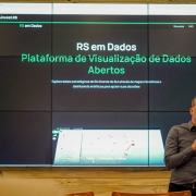A imagem mostra uma apresentação em um ambiente interno, provavelmente um auditório ou sala de conferências. No centro da imagem, há uma grande tela dividida em vários painéis exibindo a plataforma "RS em Dados", com o texto:

"RS em Dados – Plataforma de Visualização de Dados Abertos. Explore dados estratégicos do Rio Grande do Sul através de mapas interativos e dashboards analíticos para apoiar suas decisões."

À direita da imagem, está um homem careca, vestindo camisa cinza, segurando um microfone enquanto fala. Ele parece estar apresentando ou explicando a plataforma. À esquerda, de costas, está uma mulher assistindo à apresentação.

O ambiente sugere uma ocasião formal de lançamento ou demonstração de um sistema digital governamental. A interface da plataforma exibida na tela tem fundo escuro com textos e elementos gráficos em verde e branco.
