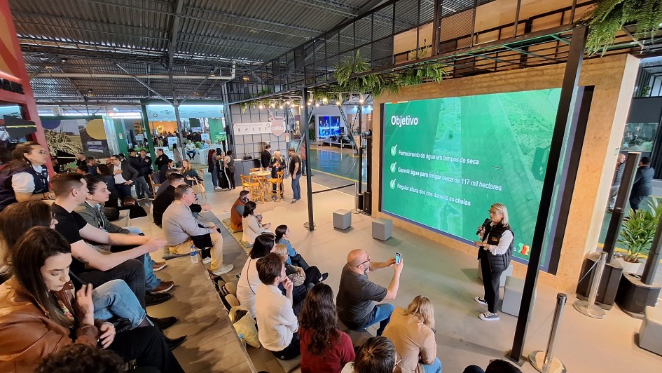 Imagem mostra uma palestra realizada em espaço fechado, com arquibancada ocupada por diversas pessoas que acompanham a apresentação. No palco, uma mulher segura microfone e fala diante de um grande telão verde que exibe o título “Objetivo” e três tópicos: fornecimento de água em tempos de seca; garantia de irrigação para cerca de 117 mil hectares; e regulação da altura dos rios durante as cheias. O ambiente é moderno, com estrutura metálica, iluminação artificial e estandes ao fundo.