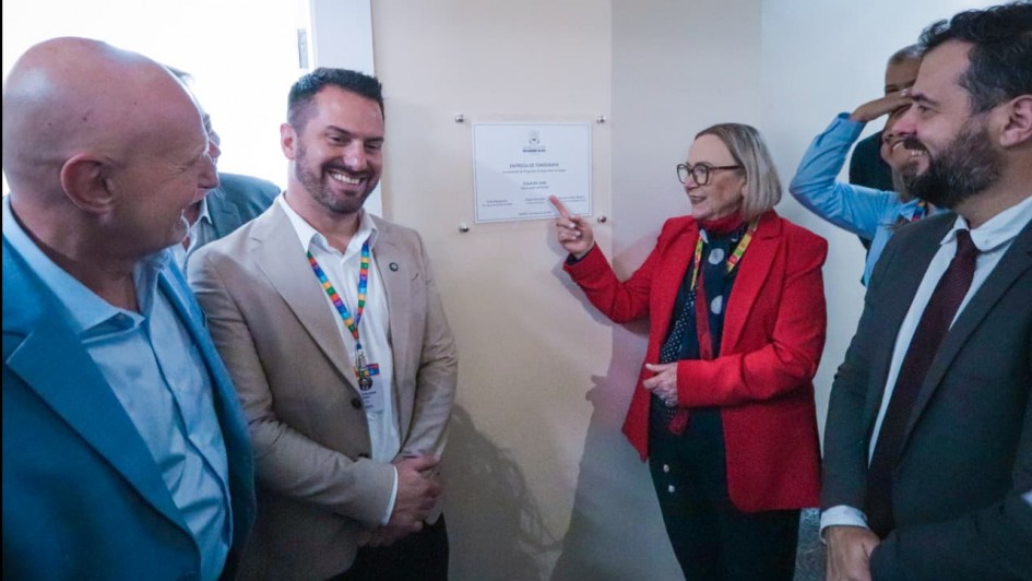 Foto mostra a secretária Arita Bergamann apontando para um placa branca na inauguração. Há outras autoridades engravatadas que sorriem.