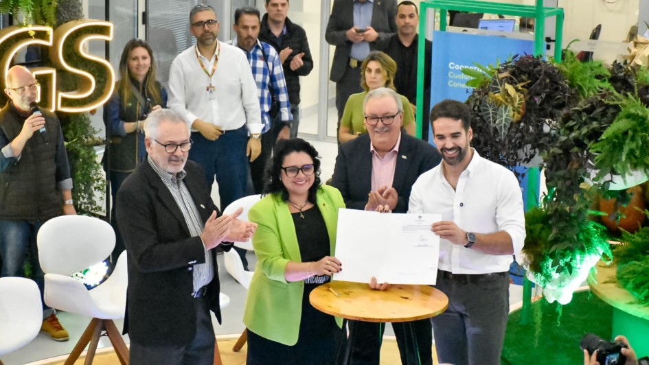 A imagem mostra, em primeiro plano, cinco pessoas em torno de uma mesa redonda de madeira clara. Quatro delas — três homens e uma mulher — estão de pé e aplaudindo, enquanto um quinto homem, de camisa social branca, segura um documento com as duas mãos. Há um público e outros indivíduos ao fundo, em um cenário que se assemelha a um evento. No canto superior esquerdo, um letreiro luminoso exibe a sigla "GS".