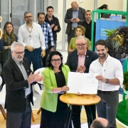A imagem mostra, em primeiro plano, cinco pessoas em torno de uma mesa redonda de madeira clara. Quatro delas — três homens e uma mulher — estão de pé e aplaudindo, enquanto um quinto homem, de camisa social branca, segura um documento com as duas mãos. Há um público e outros indivíduos ao fundo, em um cenário que se assemelha a um evento. No canto superior esquerdo, um letreiro luminoso exibe a sigla "GS".