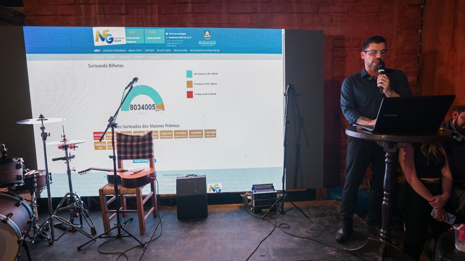 Um homem está de pé e fala ao microfone no canto direito da imagem. Ao fundo, há um telão com dados sobre os sorteados do Nota Fiscal Gaúcha de agosto. No palco, também há uma cadeira e instrumentos musicais.