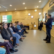 Foto mostra palestrante numa sala com paredes em madeira clara, em pé, com um microfone na mão. Ele está diante de uma plateia que o acompanha atentamente.