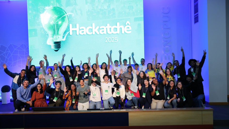 Um grupo de pessoas est&aacute; reunido em cima de um palco com as m&atilde;os para cima, posando para a foto. Ao fundo, no tel&atilde;o, l&ecirc;-se "HackaTch&ecirc; 2025". 