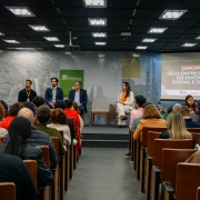 Fotografia de um auditório com cadeiras ocupadas por diversas pessoas, todas voltadas para o palco. No palco, há uma mesa com quatro pessoas sentadas — duas mulheres e dois homens — e, ao lado, uma mulher em pé, falando ao microfone em um púlpito de madeira. No fundo, um telão exibe a mensagem: “Lançamento – Selo Empresa Parceira da Diversidade Sexual e de Gênero”. Atrás do palco, há um painel com imagens e o logotipo do Governo do Estado do Rio Grande do Sul. O ambiente é iluminado e transmite o clima de um evento formal de lançamento.