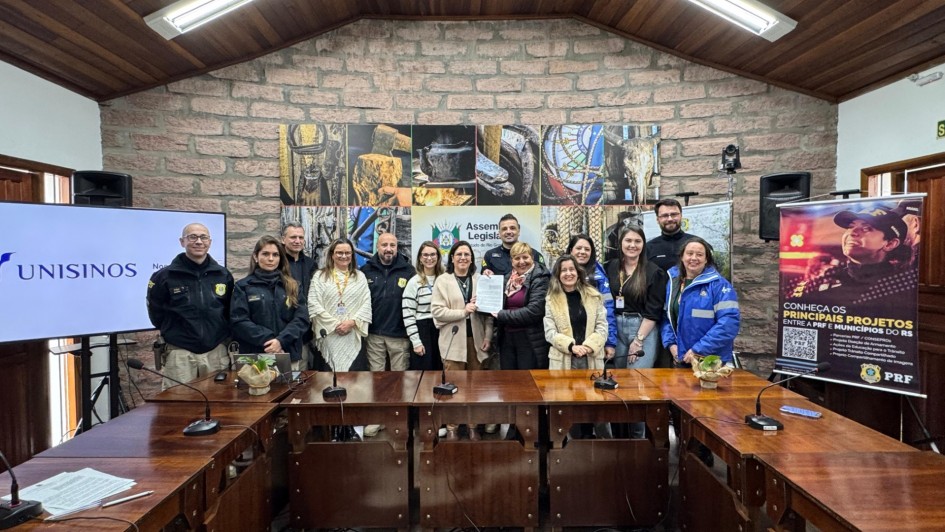 A foto mostra um grupo de aproximadamente 15 pessoas em pé atrás de uma grande mesa em formato de U. Todos parecem estar participando de um evento de assinatura de parceria. No lado esquerdo da imagem, há um telão com o logo da UNISINOS. Na parede ao fundo, há quadros com imagens rústicas e um banner colorido da PRF com o título "CONHEÇA OS PRINCIPAIS PROJETOS". A sala tem paredes de pedra e madeira no teto, com iluminação artificial forte. As pessoas vestem uma mistura de roupas casuais e uniformes, incluindo alguns da Polícia Rodoviária Federal (PRF). Uma mulher no centro do grupo está segurando um documento, possivelmente um acordo ou termo de parceria.