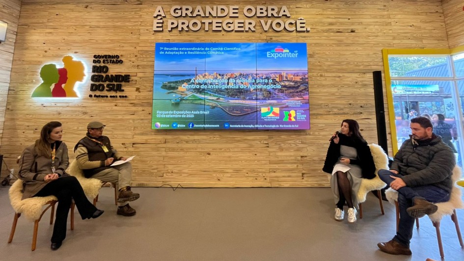 foto mostra quatro debatedores do painel sobre ciência. Eles falam sentados em cadeiras com pelegos claros e estão em frente a uma parede de madeira clara onde há um telão e há uma frase "A grende obra é proteger você" e a logomarca do RS.