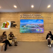 foto mostra quatro debatedores do painel sobre ciência. Eles falam sentados em cadeiras com pelegos claros e estão em frente a uma parede de madeira clara onde há um telão e há uma frase "A grende obra é proteger você" e a logomarca do RS.