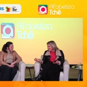 Imagem de um evento do programa Alfabetiza Tchê. Duas mulheres estão sentadas em poltronas no palco, conversando entre si. À esquerda, uma mesa lateral com garrafa de água e copo. No canto inferior direito da tela, aparece uma janela com uma intérprete de Libras usando fones de ouvido e óculos. O fundo do palco é amarelo, com os logotipos da Undime-RS, Famurs e do Governo do Estado do Rio Grande do Sul – Secretaria da Educação, além da identidade visual do programa Alfabetiza Tchê.