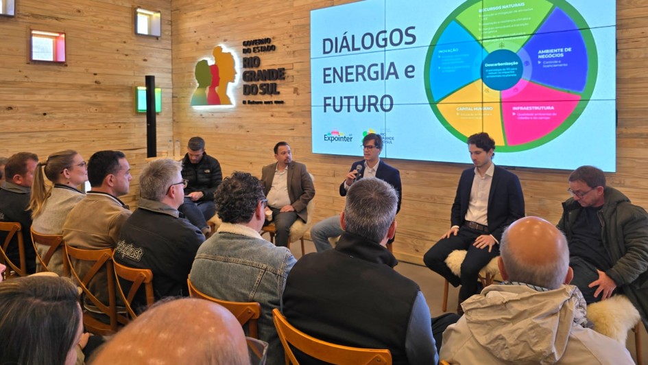 Foto mostra cinco painelistas sentados diante da plateia. O homem do centro está falando ao microfone. Atrás dele há um telão onde está escrito Diálogos Energia e Futuro e um gráfico em circulo, não legível na foto. Ao lado do telão, a logomarca do RS. 