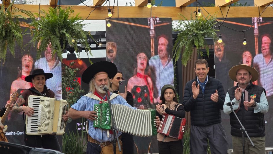 A foto captura um momento vibrante durante a abertura oficial da Campanha Ganha-Ganha Farroupilha na 48ª Expointer. Um grupo de artistas, vestindo trajes típicos gaúchos, se apresenta com acordeões e tambores, entoando músicas e danças que celebram a cultura do Rio Grande do Sul. Ao fundo, um telão exibe imagens de apresentações anteriores, reforçando o clima festivo do evento. A alegria e o orgulho das tradições sul-rio-grandenses são evidentes em cada sorriso e movimento.