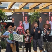 A foto captura um momento vibrante durante a abertura oficial da Campanha Ganha-Ganha Farroupilha na 48ª Expointer. Um grupo de artistas, vestindo trajes típicos gaúchos, se apresenta com acordeões e tambores, entoando músicas e danças que celebram a cultura do Rio Grande do Sul. Ao fundo, um telão exibe imagens de apresentações anteriores, reforçando o clima festivo do evento. A alegria e o orgulho das tradições sul-rio-grandenses são evidentes em cada sorriso e movimento.