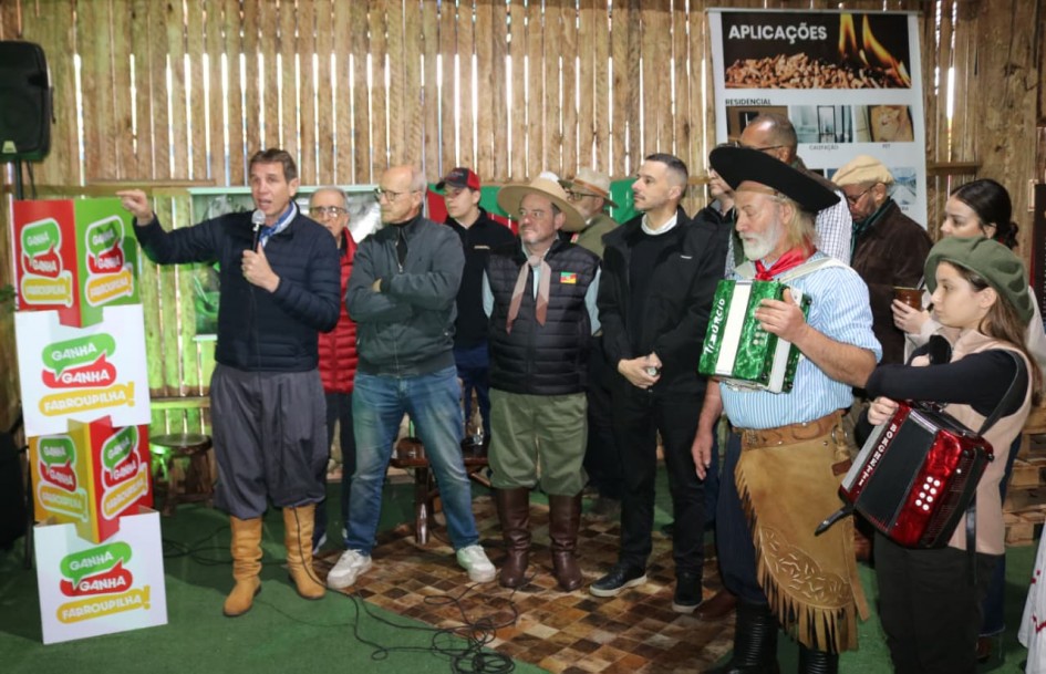 Nesta imagem, a atenção se volta para um palanque onde autoridades e artistas interagem durante a abertura oficial da Campanha Ganha-Ganha Farroupilha na 48ª Expointer. Um homem, segurando um microfone, discursa para a plateia, enquanto outros presentes, incluindo um artista com traje tradicional gaúcho e acordeão, demonstram atenção. Pilhas de caixas com a identidade visual da campanha "Ganha-Ganha Farroupilha" estão expostas, destacando o lançamento e o apoio a iniciativas locais.