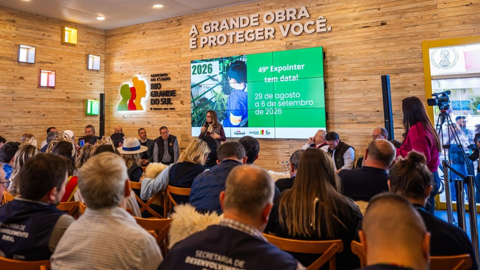 Foto mostra a coletiva da Expointer, com público sentado acompanhando apresentação. No telão, as datas de 2026. A parede do local é em madeira clara. Acima do telão a frase "A grande obra é proteger você" e a logomarca do RS. 