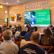 Foto mostra a coletiva da Expointer, com público sentado acompanhando apresentação. No telão, as datas de 2026. A parede do local é em madeira clara. Acima do telão a frase "A grande obra é proteger você" e a logomarca do RS. 