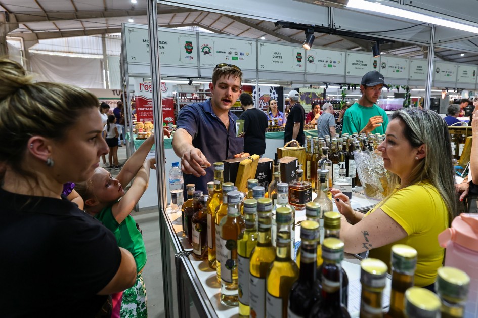 Esta fotografia de perto revela um balcão de exposição com diversos produtos, principalmente garrafas de azeite e outros itens alimentícios. Um vendedor interage com clientes, explicando os produtos. A cena destaca a presença da agricultura familiar e de pequenos produtores na Expointer, mostrando a diversidade de produtos que vão desde alimentos a bebidas. O foco está na comercialização e na valorização dos produtos locais.