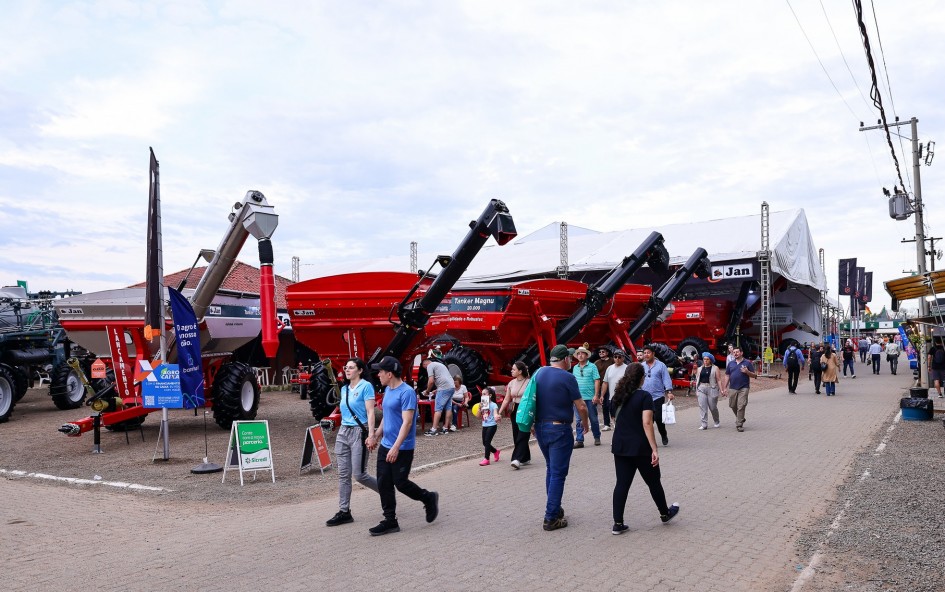 A imagem retrata uma área externa da Expointer, onde máquinas agrícolas de grande porte, como carretas e colheitadeiras, estão expostas. A cor vermelha vibrante das máquinas contrasta com o cenário mais neutro do pavilhão ao fundo. Um grupo de pessoas caminha pela área, demonstrando interesse pelos equipamentos. A presença de sinalização e a variedade de pessoas sugerem um ambiente de exposição e negócios, focado no setor agrícola.