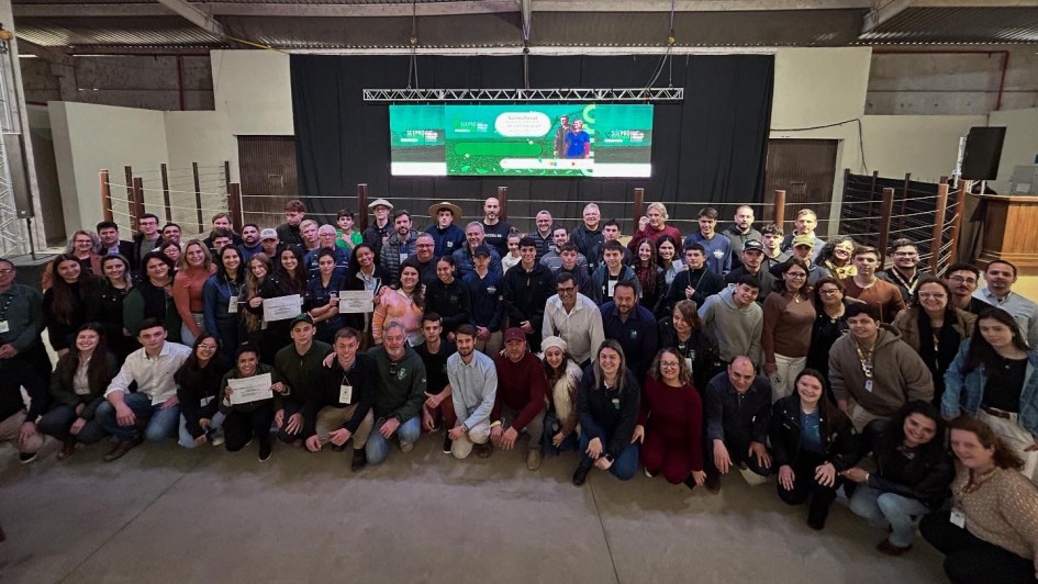 Foto posada de mais de 60 estudantes, professores e diretores em ambiente fechado. A maioria está de pé, alguns se encontram agachados à frente. Ao fundo aparece um telão.