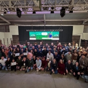 Foto posada de mais de 60 estudantes, professores e diretores em ambiente fechado. A maioria está de pé, alguns se encontram agachados à frente. Ao fundo aparece um telão.