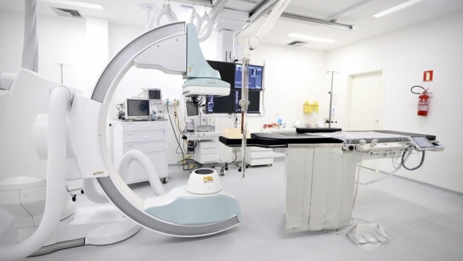 A imagem mostra uma sala de hemodinâmica em um hospital, equipada com aparelhos modernos utilizados para diagnóstico e tratamento de doenças cardiovasculares. No centro, há uma mesa de exames com monitores acoplados e, ao lado, um equipamento em formato de arco para realização de procedimentos de imagem. O ambiente é amplo, iluminado e de paredes brancas, transmitindo aspecto de assepsia. À direita, há um extintor fixado na parede, reforçando as normas de segurança do espaço hospitalar.