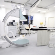A imagem mostra uma sala de hemodinâmica em um hospital, equipada com aparelhos modernos utilizados para diagnóstico e tratamento de doenças cardiovasculares. No centro, há uma mesa de exames com monitores acoplados e, ao lado, um equipamento em formato de arco para realização de procedimentos de imagem. O ambiente é amplo, iluminado e de paredes brancas, transmitindo aspecto de assepsia. À direita, há um extintor fixado na parede, reforçando as normas de segurança do espaço hospitalar.