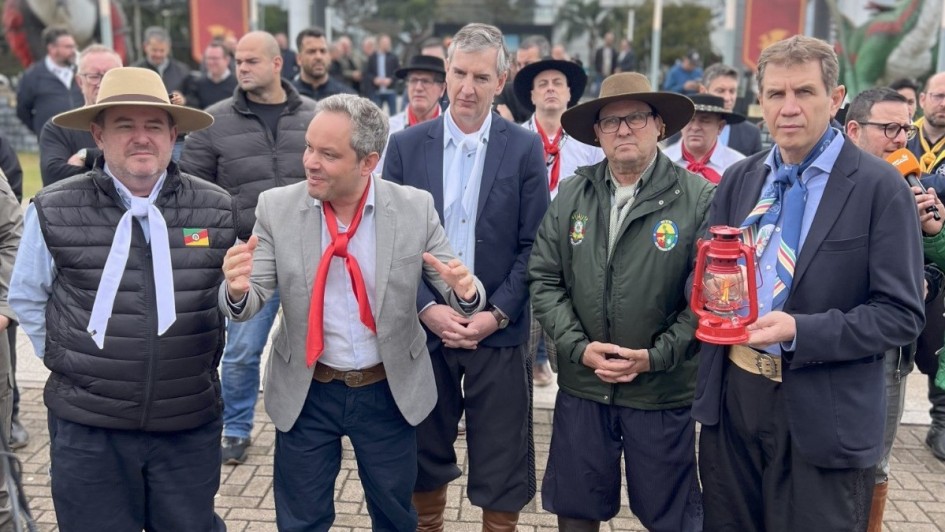 Cena mostra o secretário Ernani Polo segurando uma lamparina com a Chama Crioula, em pé à direita da foto. Ao seu lado esquerdo estão os secretários da Cultura, Eduardo Loureiro, e de Turismo, Ronaldo Santini, e dois tradicionalistas. Todos estão pilchados.