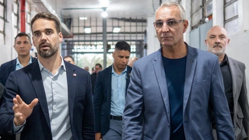 Dois homens caminham lado a lado dentro de uma unidade prisional. Ao fundo, há outros homens acompanhando. O governador está ao centro da imagem, conversando com um agente da Polícia Penal uniformizado, que caminha ao seu lado.