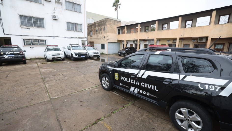 Foto de um carro de polícia preto estacionado no pátio da delegacia, à direita do quadro. Ao fundo, aparecem um prédio branco e outros carros da polícia estacionados.