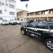 Foto de um carro de polícia preto estacionado no pátio da delegacia, à direita do quadro. Ao fundo, aparecem um prédio branco e outros carros da polícia estacionados.