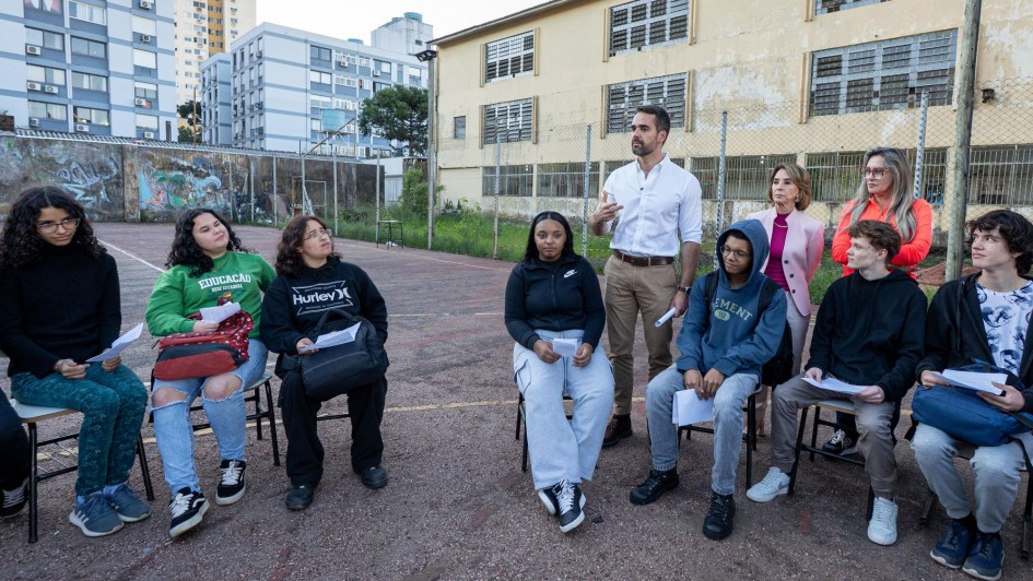 O governador Eduardo Leite está em um pátio escolar, conversando com um grupo de estudantes que estão sentados em cadeiras, em um círculo. Ele gesticula enquanto fala, indicando que está explicando algo ou ouvindo atentamente. Ao fundo, o prédio do colégio e outros edifícios residenciais compõem a paisagem.