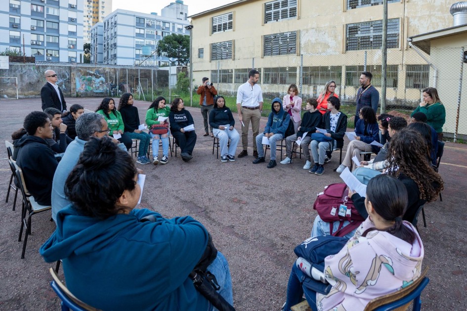 O governador Eduardo Leite está em um pátio escolar, conversando com um grupo de estudantes que estão sentados em cadeiras, em um círculo. Ele gesticula enquanto fala, indicando que está explicando algo ou ouvindo atentamente. Ao fundo, o prédio do colégio e outros edifícios residenciais compõem a paisagem.