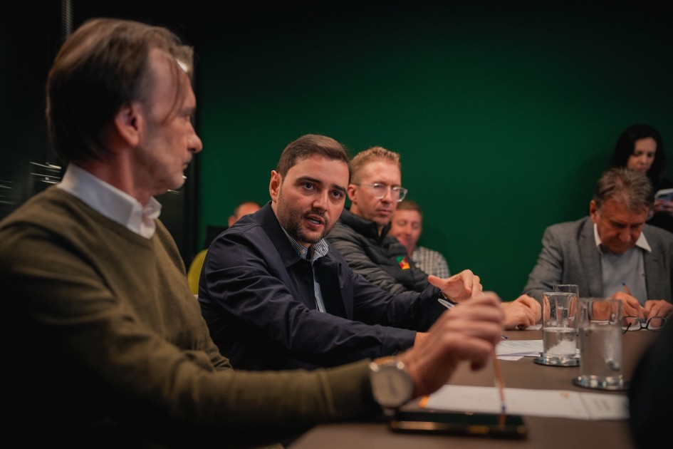Nesta foto, o vice-governador Gabriel Souza está sentado à mesa de reuniões, olhando para a pessoa que está falando à sua esquerda. Ele casaco azul escuro. A imagem foca em um grupo de pessoas reunidas em torno de uma mesa, em um ambiente de trabalho formal. Ao fundo, uma parede verde escuro.