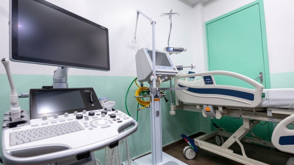 A imagem mostra um quarto hospitalar moderno, equipado com cama hospitalar ajustável de metal e plástico, com grades laterais e rodízios. Ao lado, há equipamentos médicos avançados, incluindo monitor multiparamétrico, respirador mecânico e máquina de ultrassom com teclado e tela de alta definição. As paredes em branco e verde-claro, junto da porta verde, reforçam a sensação de limpeza e organização. O espaço é iluminado artificialmente e reflete um ambiente preparado para cuidados intensivos.