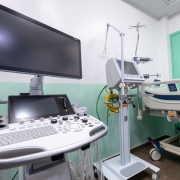 A imagem mostra um quarto hospitalar moderno, equipado com cama hospitalar ajustável de metal e plástico, com grades laterais e rodízios. Ao lado, há equipamentos médicos avançados, incluindo monitor multiparamétrico, respirador mecânico e máquina de ultrassom com teclado e tela de alta definição. As paredes em branco e verde-claro, junto da porta verde, reforçam a sensação de limpeza e organização. O espaço é iluminado artificialmente e reflete um ambiente preparado para cuidados intensivos.