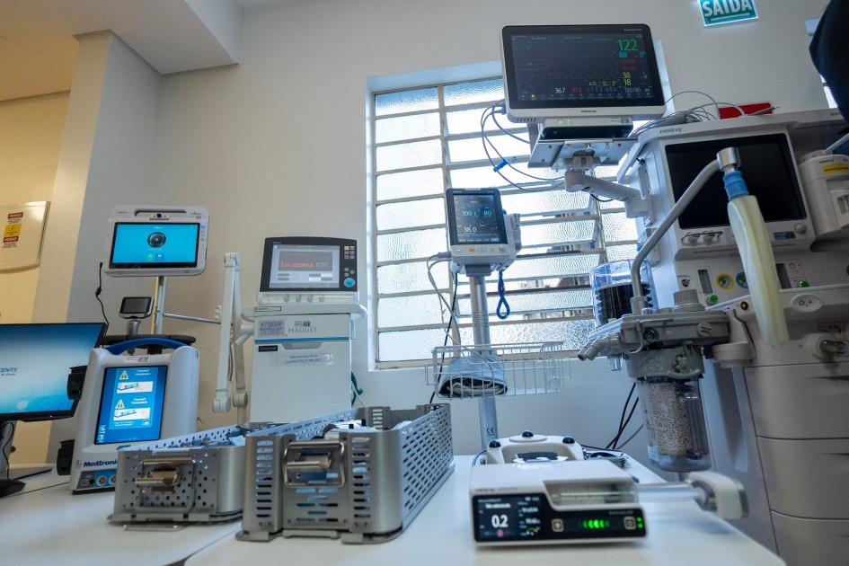 A foto registra uma sala hospitalar repleta de equipamentos médicos de última geração, organizados sobre bancadas e suportes. É possível ver monitores de sinais vitais ligados, respiradores, bombas de infusão e aparelhos de anestesia, todos conectados por cabos e tubos. A luz natural entra pela janela com grades, iluminando o espaço e destacando o caráter técnico do ambiente. A cena transmite modernidade e alta capacidade de suporte em terapias intensivas, reforçando a ideia de um hospital estruturado.