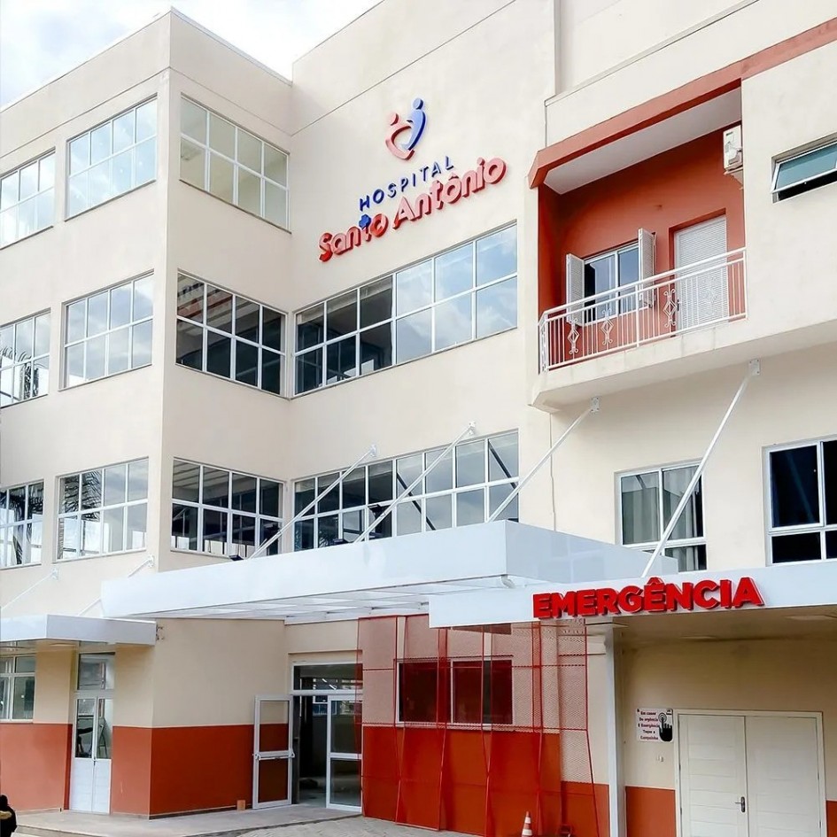 A imagem mostra a fachada principal de um hospital de médio porte, com três andares, paredes claras e janelas amplas de vidro. Na entrada principal, há uma estrutura branca moderna e, logo abaixo, a placa vermelha com a palavra “Emergência”, indicando o setor de pronto atendimento.