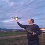 A foto mostra um homem em um campo aberto, no fim da tarde, segurando um controle remoto de drone com a mão esquerda enquanto lança o equipamento com a mão direita. O drone, iluminado por luzes verdes, está em voo próximo à sua mão. O céu está parcialmente nublado, com tons azulados e rosados, enquanto ao fundo aparecem cercas, casas rurais, veículos estacionados e a silhueta de montanhas.