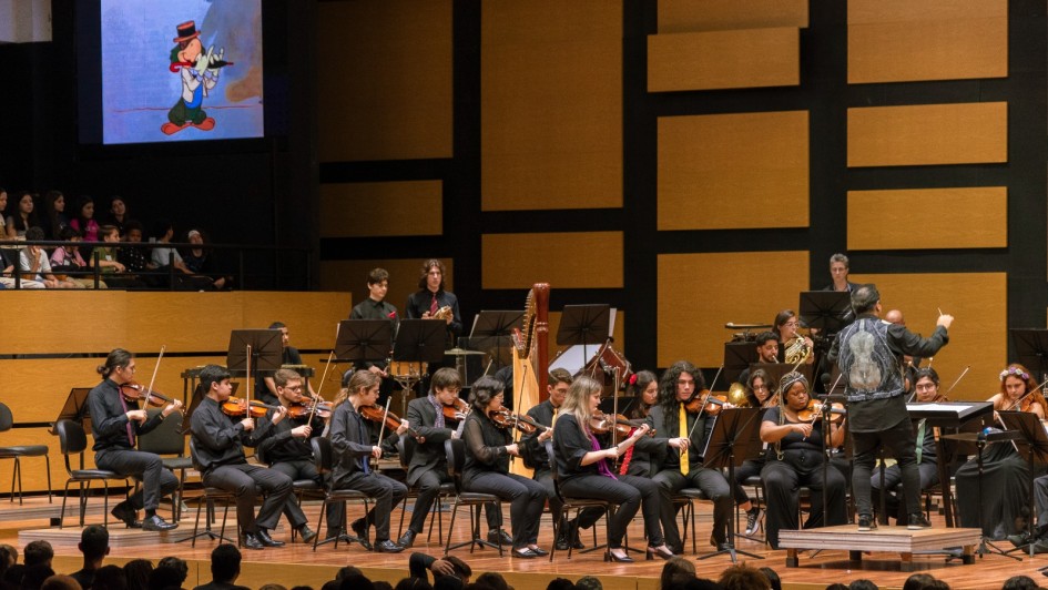 Esta é uma imagem de uma orquestra em uma apresentação, provavelmente em uma sala de concertos. A orquestra está composta principalmente por músicos tocando violinos e outros instrumentos, como harpa e trompa. O maestro está de costas para a câmera, conduzindo a orquestra.

No fundo, à esquerda, há uma tela com uma imagem animada de um personagem que parece ser o Pato Donald, vestido com chapéu e casaco, tocando um instrumento de sopro. A plateia está sentada em frente ao palco, assistindo ao concerto.

A sala tem painéis acústicos retangulares de madeira nas paredes, provavelmente para melhorar a qualidade sonora da apresentação. Os músicos estão vestidos com roupas pretas, algumas com detalhes em vermelho ou amarelo, sugerindo um código de vestimenta formal ou uniforme para o evento.