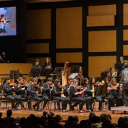 Esta é uma imagem de uma orquestra em uma apresentação, provavelmente em uma sala de concertos. A orquestra está composta principalmente por músicos tocando violinos e outros instrumentos, como harpa e trompa. O maestro está de costas para a câmera, conduzindo a orquestra.

No fundo, à esquerda, há uma tela com uma imagem animada de um personagem que parece ser o Pato Donald, vestido com chapéu e casaco, tocando um instrumento de sopro. A plateia está sentada em frente ao palco, assistindo ao concerto.

A sala tem painéis acústicos retangulares de madeira nas paredes, provavelmente para melhorar a qualidade sonora da apresentação. Os músicos estão vestidos com roupas pretas, algumas com detalhes em vermelho ou amarelo, sugerindo um código de vestimenta formal ou uniforme para o evento.