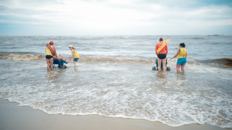 Foto em ambiente aberto do mar, com duas pessoas em cadeiras híbridas entrando na água, monitoradas por dois guarda-vidas. Elas aparecem de costas.