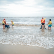 Foto em ambiente aberto do mar, com duas pessoas em cadeiras híbridas entrando na água, monitoradas por dois guarda-vidas. Elas aparecem de costas.