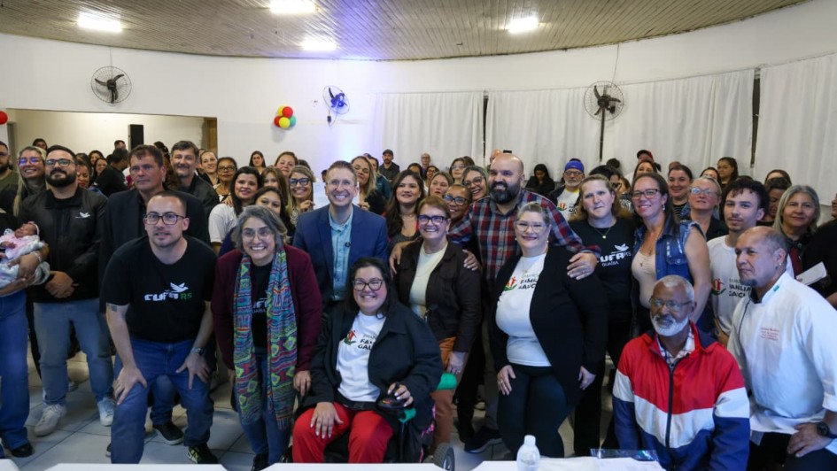 Descrição da imagem: Um grupo numeroso de pessoas está reunido em um salão iluminado, posando para uma foto coletiva. Na frente, algumas pessoas estão sentadas e outras de pé, incluindo mulheres e homens de diferentes idades e estilos. Algumas usam camisetas com os logos “Clube RS” e “Famílias Gaúchas”, sugerindo participação em um evento comunitário ou institucional. Há também uma mulher em cadeira de rodas no centro da primeira fila, sorridente. Ao fundo, outras pessoas formam uma segunda e terceira fileira, todas olhando para a câmera. O ambiente tem paredes brancas, cortinas e ventiladores fixados, além de algumas bexigas coloridas na decoração lateral. O clima é de confraternização e inclusão.