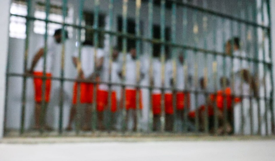 Fotografia desfocada do interior de uma cela de prisão, vista através das grades metálicas na frente. Dentro do espaço, várias pessoas estão em pé, usando camisetas brancas e bermudas laranja. A cena transmite a ideia de confinamento coletivo, com os presos posicionados lado a lado no ambiente iluminado.