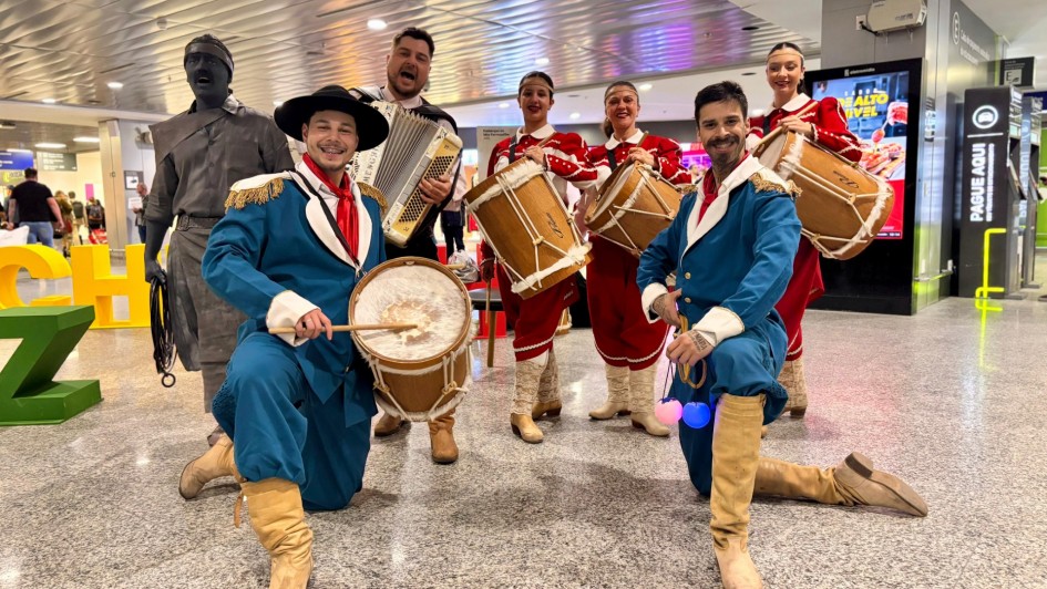 A imagem mostra um grupo de artistas trajando roupas típicas gaúchas em apresentação cultural dentro do Aeroporto Salgado Filho. Dois homens vestidos de bombacha azul, lenço vermelho e botas seguram tambores e estão ajoelhados em destaque. Atrás deles, quatro mulheres de vestido vermelho tocam instrumentos, e um homem toca acordeom. Ao fundo, um artista caracterizado como estátua humana também participa da cena. O ambiente moderno do saguão contrasta com a tradição, criando clima festivo de celebração do Mês Farroupilha.