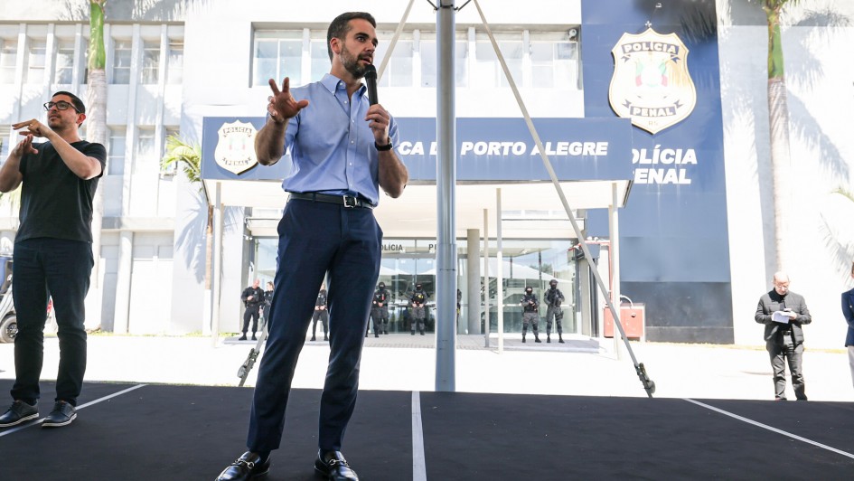 Um homem em pé sobre um palco ao ar livre fala ao microfone, usando camisa azul clara de manga comprida dobrada até os cotovelos, calça social azul-marinho e sapatos pretos. Ele gesticula com a mão esquerda enquanto fala. À esquerda da imagem, outro homem faz interpretação em Libras, vestindo camiseta preta e óculos. Ao fundo, aparece a fachada de um prédio institucional com letreiros que indicam "Polícia Penal" e "Porto Alegre", além de brasões. Em frente ao prédio, alguns policiais fardados permanecem em posição de guarda. Outro homem, à direita da foto, observa anotações em um caderno. O cenário é iluminado pela luz do dia e há palmeiras ao redor do edifício.