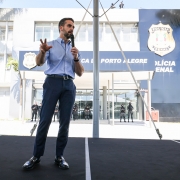 Um homem em pé sobre um palco ao ar livre fala ao microfone, usando camisa azul clara de manga comprida dobrada até os cotovelos, calça social azul-marinho e sapatos pretos. Ele gesticula com a mão esquerda enquanto fala. À esquerda da imagem, outro homem faz interpretação em Libras, vestindo camiseta preta e óculos. Ao fundo, aparece a fachada de um prédio institucional com letreiros que indicam "Polícia Penal" e "Porto Alegre", além de brasões. Em frente ao prédio, alguns policiais fardados permanecem em posição de guarda. Outro homem, à direita da foto, observa anotações em um caderno. O cenário é iluminado pela luz do dia e há palmeiras ao redor do edifício.