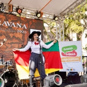 A foto mostra apresentação no palco da Caravana Farroupilha 2025, instalado ao ar livre no centro de Porto Alegre. Uma mulher com trajes gaúchos tradicionais, chapéu preto e lenço vermelho, segura a bandeira do Rio Grande do Sul enquanto canta ao microfone. Ao lado, um homem toca violão, e ao fundo há bateria e instrumentos. O painel atrás exibe “Caravana Farroupilha 2025”. 